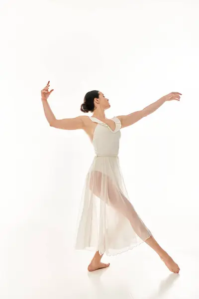 Молода жінка в довгій білій сукні витончено танцює в студії на білому тлі. — стокове фото