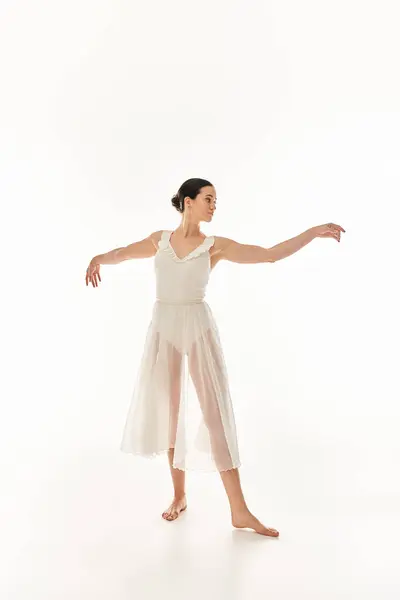 Молода жінка в довгій білій сукні стоїть з руками, витягнутими у витонченій танцювальній позі на білому фоні студії. — стокове фото