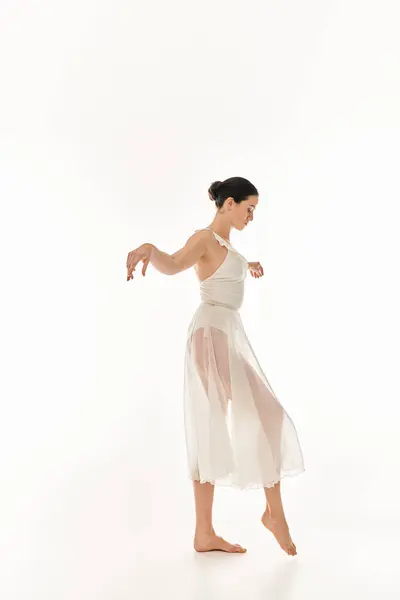 Una mujer joven baila con gracia en un vestido blanco que fluye sobre un fondo blanco en un entorno de estudio. - foto de stock