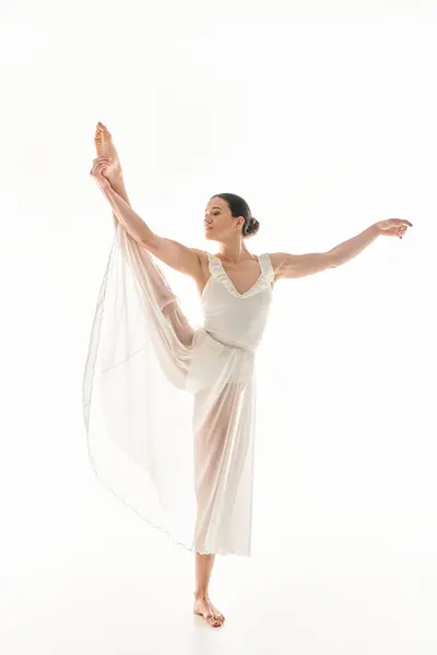 Une jeune femme danse gracieusement dans une longue robe blanche sur fond blanc dans un décor de studio. — Photo de stock