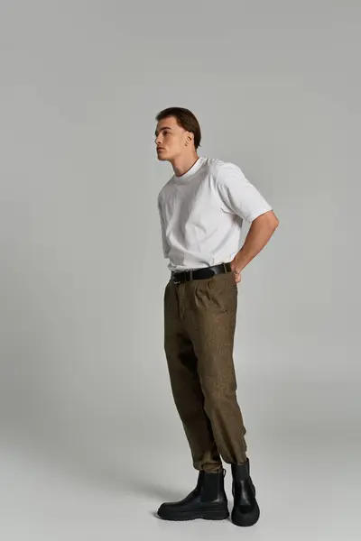 Stylish young man in t shirt and brown pants posing attractively on gray backdrop and looking away — Stock Photo