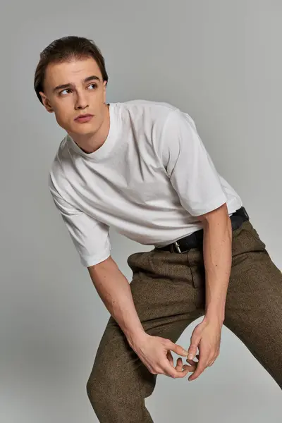 Handsome young man in t shirt and brown pants posing attractively on gray backdrop and looking away — Stock Photo