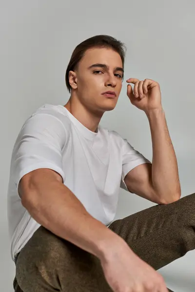 Atractivo joven pulido en ropa elegante sentado sobre fondo gris y mirando a la cámara - foto de stock
