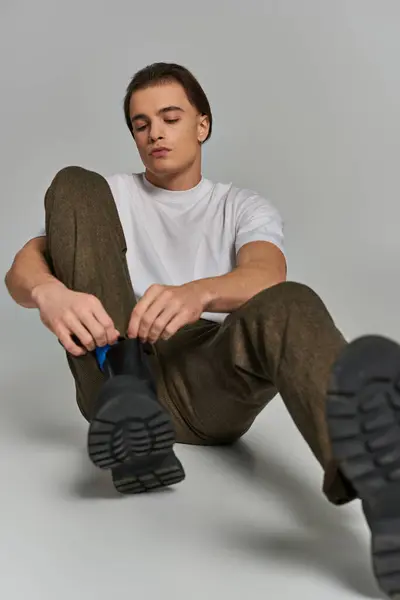 Atractivo modelo masculino debonair en pantalones marrones sentado y atando sus cordones sobre fondo gris - foto de stock