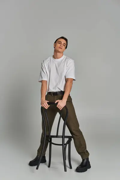 Séduisant jeune homme en tenue tendance assis attrayant sur la chaise et regardant loin sur fond gris — Photo de stock