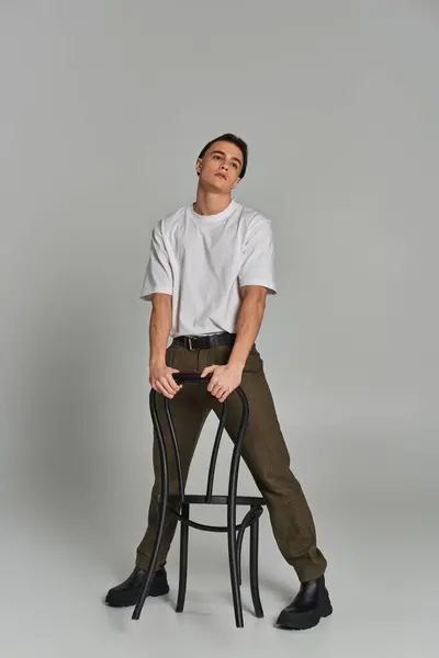 Alluring young man in trendy attire sitting attractively on chair and looking away on gray backdrop — Stock Photo