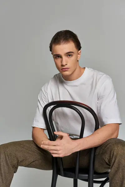 Handsome trendy man in debonair attire sitting on chair and looking at camera on gray backdrop — Stock Photo