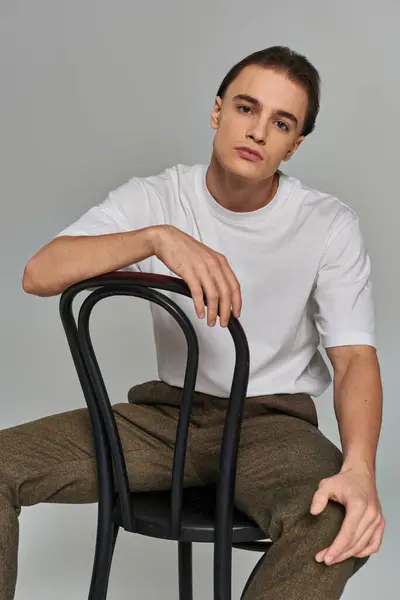 Handsome trendy man in debonair attire sitting on chair and looking at camera on gray backdrop — Stock Photo