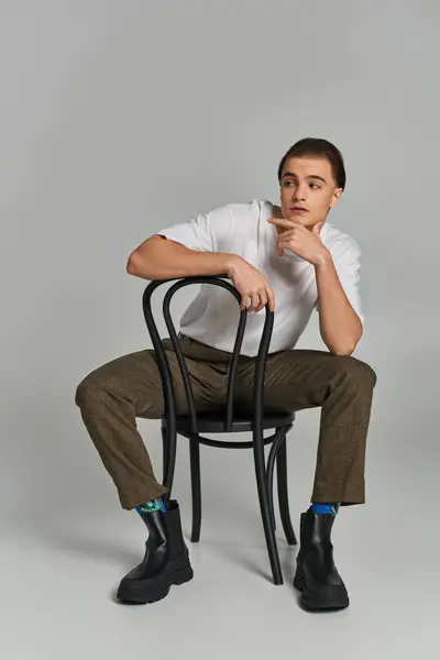 Séduisant jeune homme en tenue tendance assis attrayant sur la chaise et regardant loin sur fond gris — Photo de stock