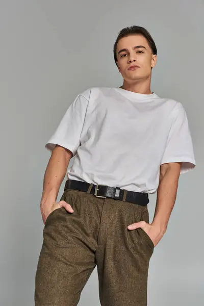 Atraente jovem modelo masculino em calças sofisticadas olhando para a câmera enquanto no fundo cinza — Fotografia de Stock