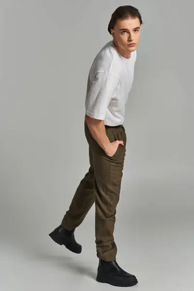 Attraente giovane uomo in t shirt e pantaloni marroni in posa attraente su sfondo grigio e guardando altrove — Foto stock