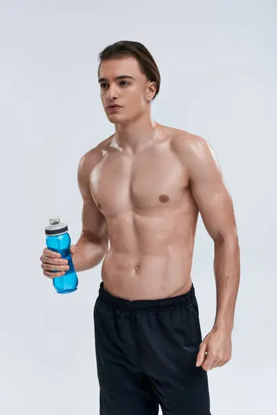Bonito sexy masculino modelo no preto calças segurando água garrafa e olhando para longe no cinza pano de fundo — Fotografia de Stock