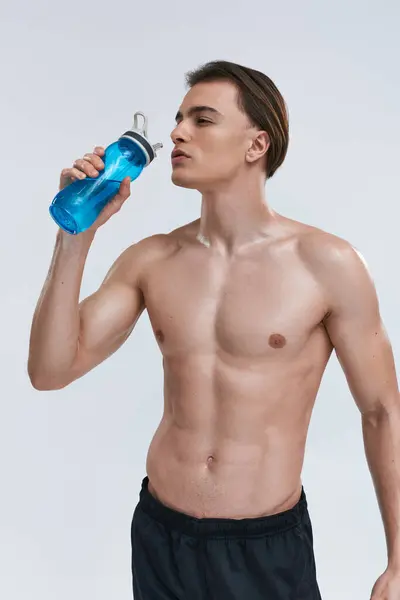 Guapo sexy sin camisa modelo masculino beber agua de botella y mirando hacia otro lado en gris telón de fondo - foto de stock