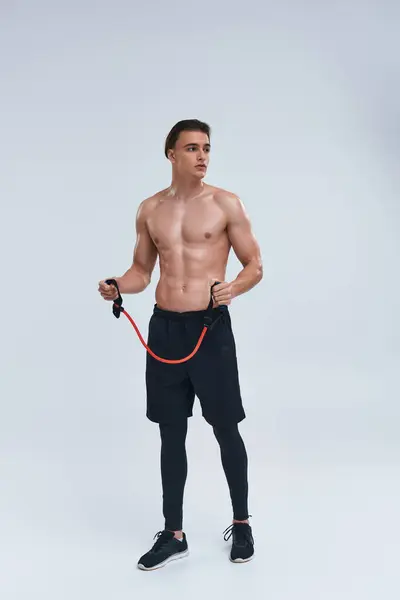 Tentador joven deportivo sin camisa en pantalones negros entrenando con expansor de fitness y mirando hacia otro lado - foto de stock