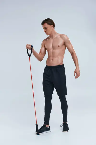 Seductor sin camisa joven deportivo en pantalones negros de entrenamiento con expansor de fitness y mirando hacia otro lado - foto de stock