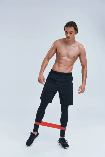 Atractivo joven deportivo en pantalones negros posando en topless con banda de resistencia y mirando hacia otro lado - foto de stock