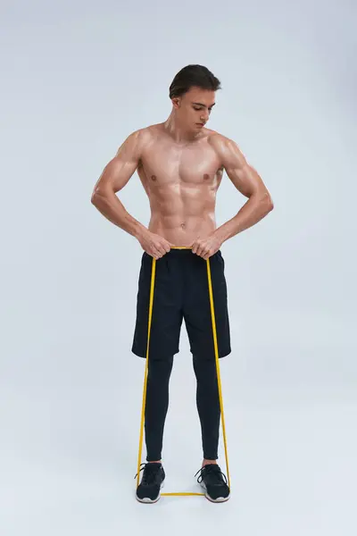 Séduisant jeune homme athlétique en pantalon noir posant seins nus avec bande de résistance et détournant les yeux — Photo de stock