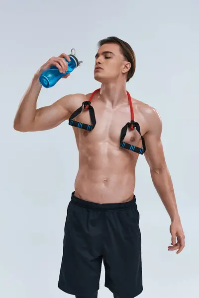 Tentador hombre deportivo sin camisa en pantalones negros bebiendo agua con expansor de fitness en su cuello - foto de stock