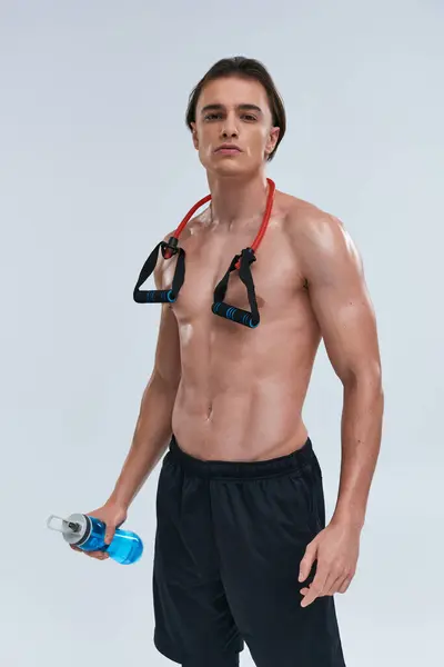 Athlétique sexy jeune homme posant seins nus avec bouteille d'eau et extenseur de fitness et en regardant la caméra — Photo de stock