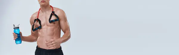 Vista recortada de hombre deportivo en pantalones negros posando en topless con botella y expansor de fitness, pancarta - foto de stock