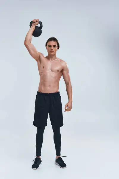 Atraente atlético sem camisa homem exercitando com kettlebell e olhando para a câmera no fundo cinza — Fotografia de Stock