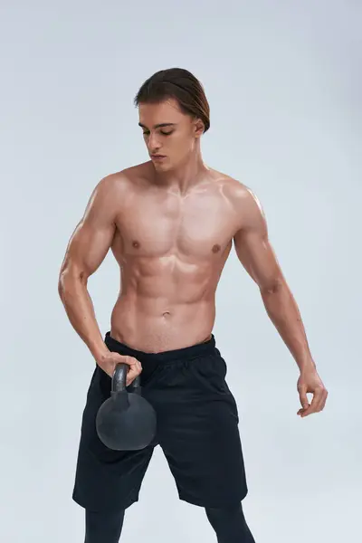 Alluring athletic young man in black sporty pants exercising with kettlebell on gray background — Stock Photo