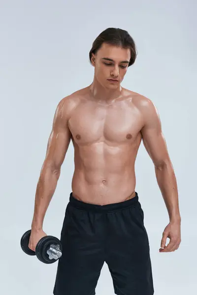 Atractivo joven sin camisa hombre en negro pantalones cómodos ejercicio con mancuerna y mirando hacia otro lado - foto de stock