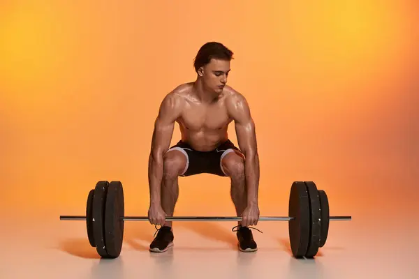 Alluring shirtless muscular man in black pants exercising with barbell on vibrant orange backdrop — Stock Photo