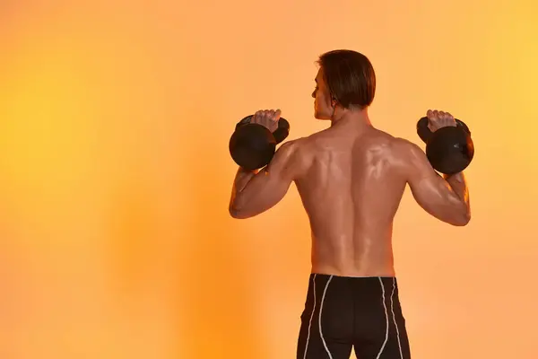 Back view of young muscular man posing topless while exercising with kettlebells on orange backdrop — Stock Photo