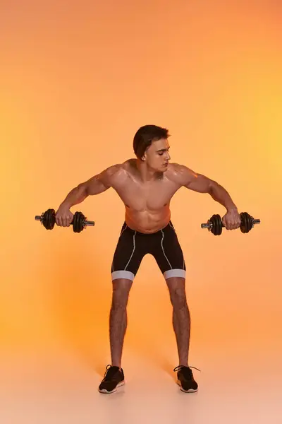 Attraente uomo muscoloso senza maglietta in pantaloncini sportivi neri formazione con manubri su sfondo vivido — Foto stock