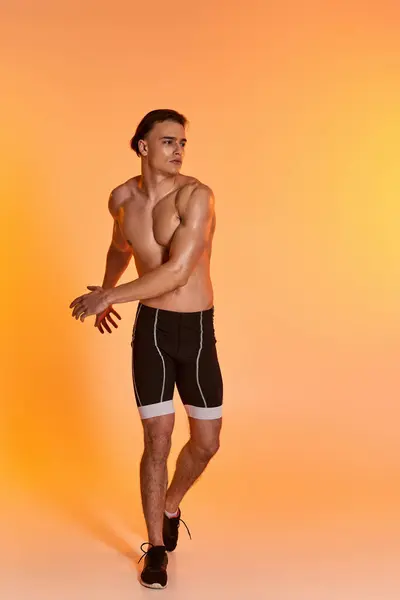 Hombre guapo sin camisa en pantalones cortos negros haciendo ejercicio activamente y mirando hacia otro lado en el fondo naranja - foto de stock