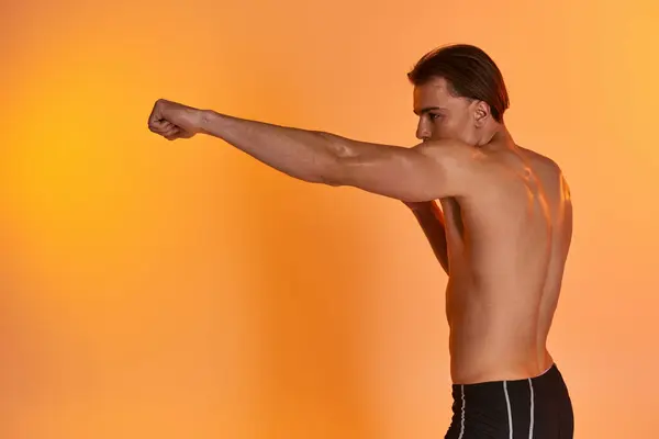 Alluring shirtless sexy man in black sporty shorts boxing actively on vibrant orange background — Stock Photo