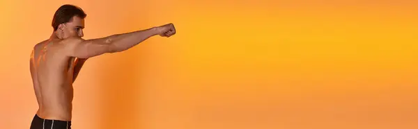 Alluring shirtless sexy man in black shorts boxing actively on vibrant orange background, banner — Stock Photo