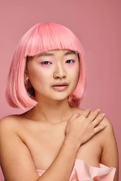 Encantadora mujer asiática con pelo rosa y maquillaje tocando su hombro con la mano sobre fondo vibrante - foto de stock