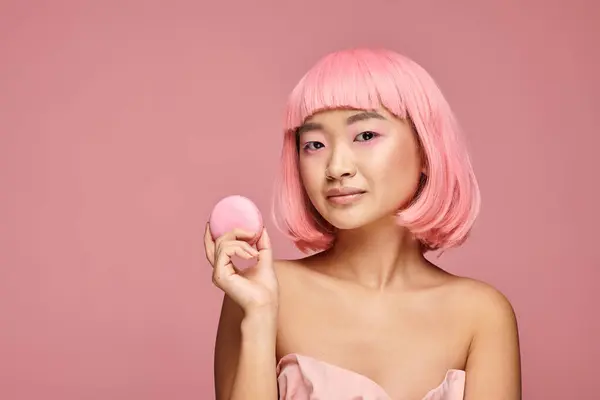 Bastante asiático joven mujer con rosa pelo y maquillaje celebración macarrón con mano en vibrante fondo - foto de stock