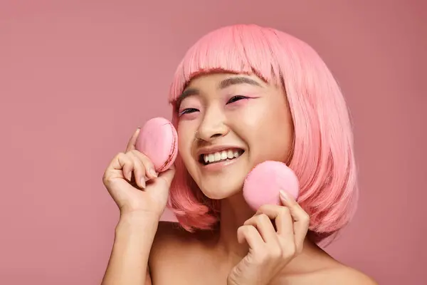 Feliz menina asiática com o cabelo rosa rindo e segurando macaroons com a mão em fundo vibrante — Fotografia de Stock
