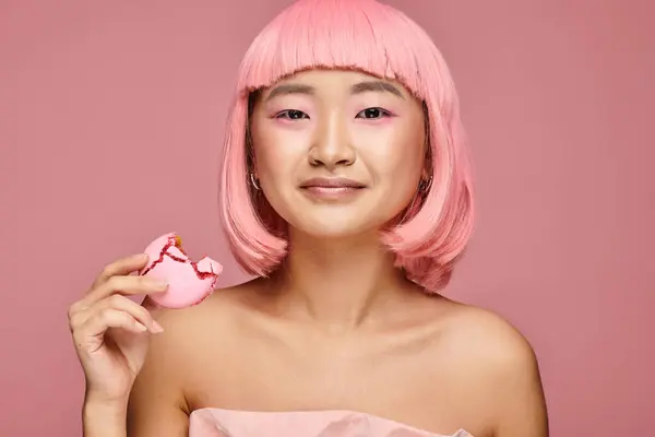 Attrayant asiatique femme avec rose cheveux et maquillage posant avec morsure de macaron sur fond vibrant — Photo de stock