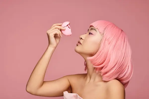 Профиль милой азиатской девушки с розовыми волосами, позирующими укус макарона на ярком фоне — стоковое фото