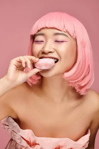 Atractivo asiático joven mujer con rosa pelo y cerrado ojos comer mochi contra vibrante fondo - foto de stock