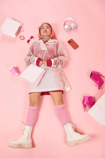 Encantador mulher asiática com cabelo rosa em roupa elegante deitado com compras em fundo vibrante — Fotografia de Stock