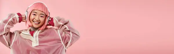 Banner de chica asiática en 20s con pelo rosa escuchando música con auriculares en un fondo vibrante - foto de stock
