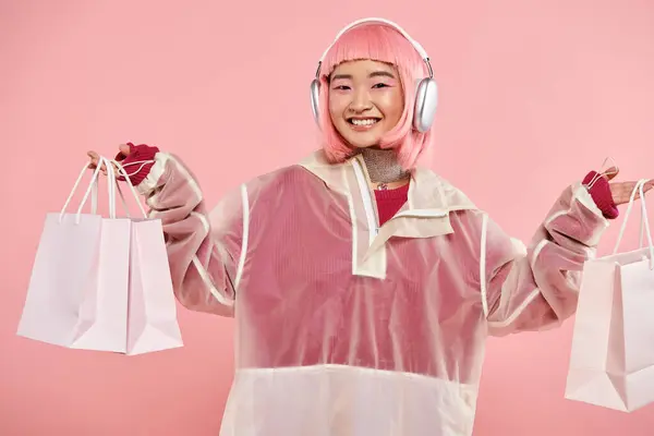 Atraente mulher asiática com cabelo rosa e sacos de compras em fones de ouvido no fundo vibrante — Fotografia de Stock