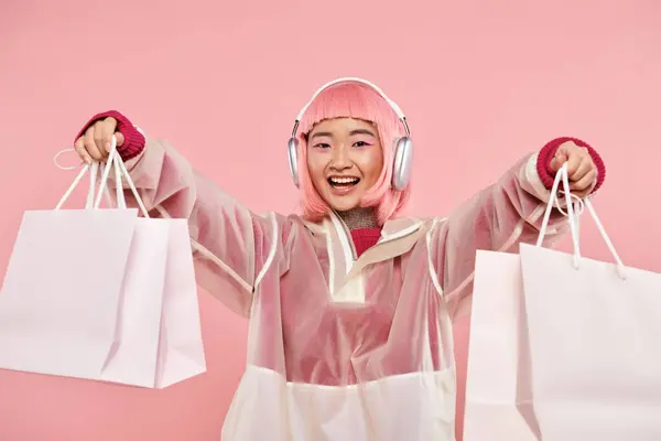 Femme asiatique gaie avec des cheveux roses et des sacs à provisions dans des écouteurs en arrière-plan vibrant — Photo de stock