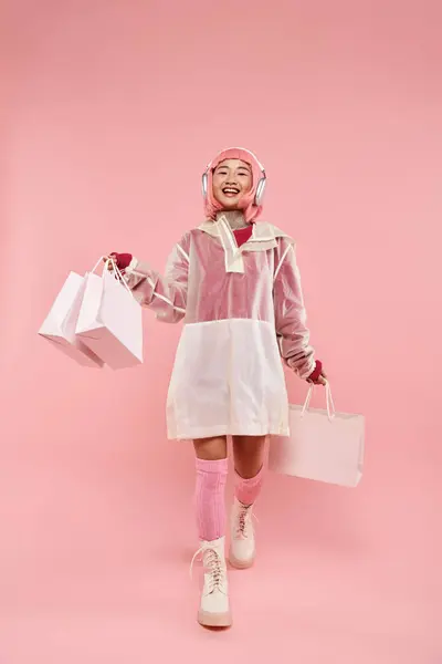Feliz menina asiática com cabelo rosa e sacos de compras em fones de ouvido andando sobre fundo vibrante — Fotografia de Stock