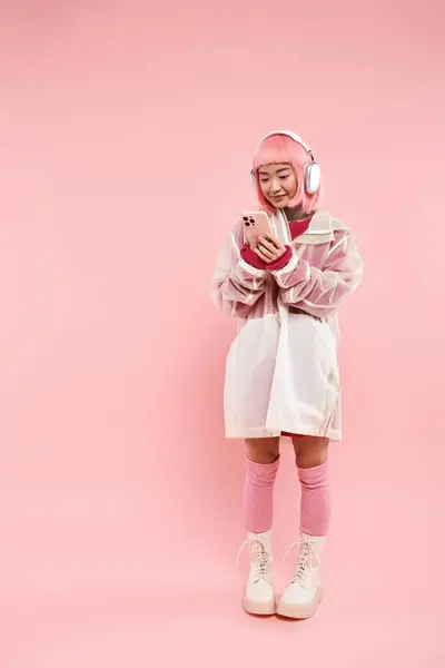 Atractiva mujer asiática con pelo rosa en auriculares mensajes de texto en el teléfono inteligente sobre fondo vibrante - foto de stock