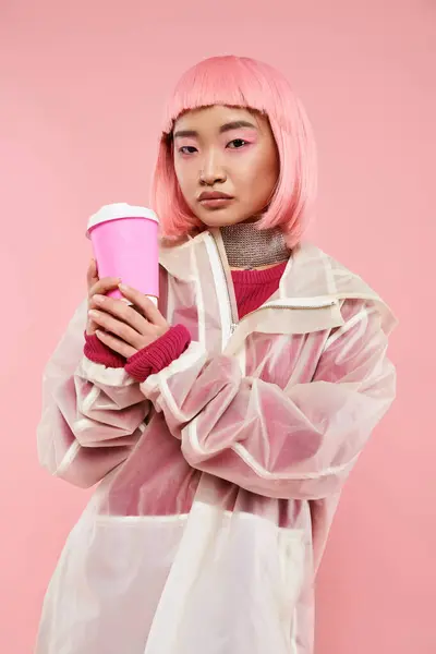 Seguro asiático joven mujer con rosa pelo posando con café taza sobre vibrante fondo - foto de stock
