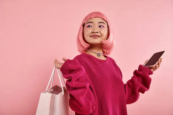 Pretty asian young woman with shopping packet and smartphone looking to side on pink background — Stock Photo