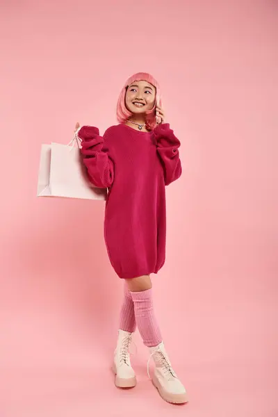 Asian woman with pink hair calling of smartphone and holding packet over shoulder on pink background — Stock Photo