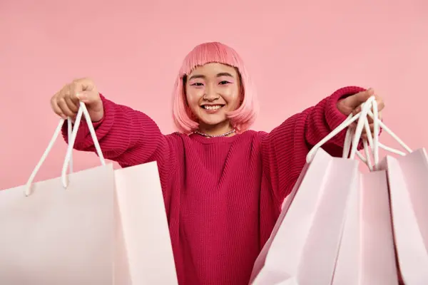 Hermosa chica joven asiática sonriendo ampliamente y sosteniendo hacia fuera a las bolsas de compras sobre fondo rosa - foto de stock