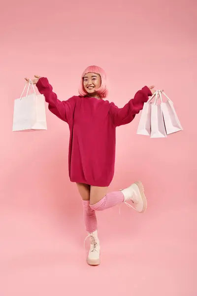 Mignonne asiatique femme dans son 20s posant avec jambe levée et tenant des sacs à provisions sur fond rose — Photo de stock